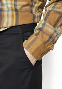 Kostkovaná hnědá a žlutá košile na knoflíky s černými kalhotami s kapsou. Košile má pruhy v modré a zelené barvě a je vyrobena z texturované látky.