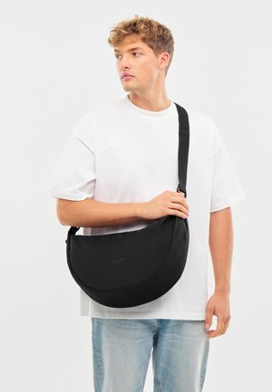 Čierna polmesiacová crossbody taška vyrobená z mäkkého textilu s nastaviteľným popruhom. Na prednej strane sa nachádza minimálne branding v debnení.