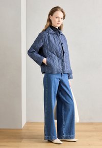 Gequilted marineblauwe jas met ruitpatroon, knoopsluiting en twee zakken, gecombineerd met lichtblauwe wijde spijkerbroek en crèmekleurige hakken.