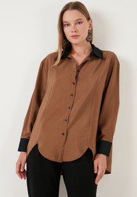 Brown long-sleeved shirt with a black collar and buttons, smooth texture, curved hem, and side slits. Worn with black pants.