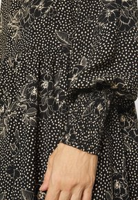 Nahaufnahme einer Hand, die in der Nähe eines schwarzen Kleides mit weißen Blumen- und Punkte-Mustern ruht, das geraffte, elastische Ärmel hat.