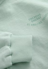 Jasnozielony rękaw bluzy z ściągaczem i nadrukiem tekstu "CREATIVE ENERGY BE AMAZING TODAY" na tkaninie.