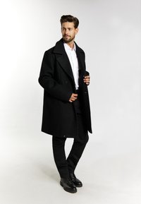 Černý vlněný kabát s širokým límcem, dvouřadové knoflíky, nošený přes bílou košili a černé kalhoty. Černé šněrovací boty.