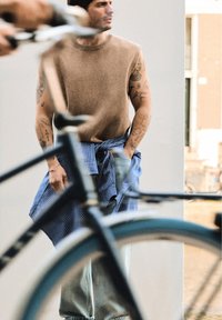 Muž s tetovanými pažemi, oblečený v hnědém svetru bez rukávů a košili uvázané v pase, stojí za černým rámem jízdního kola v městském prostředí.