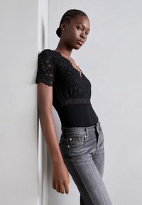 Bodysuit en dentelle noire avec des manches courtes et un décolleté en V, associé à un jean gris ajusté avec des poches et des accents métalliques.