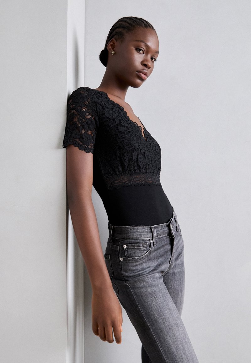 Bodysuit en dentelle noire avec des manches courtes et un décolleté en V, associé à un jean gris ajusté avec des poches et des accents métalliques.