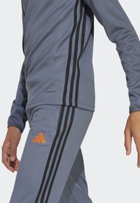 Chándal atlético gris con rayas negras a los lados, acentos en naranja en el logo y un diseño ajustado con un acabado de tela suave.