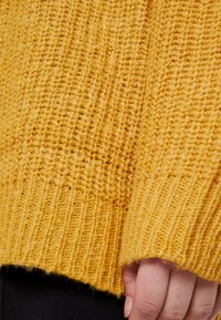Žlutý pletený svetr s texturovaným vzorem, se žebrovanými manžetami a volným střihem. Látka působí měkkým a teplým dojmem.