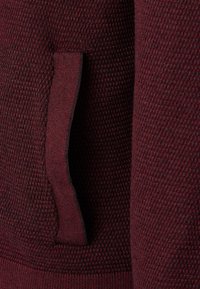 Burgundy texturerad tröja med ett vävt mönster, med en sidoficka och en ribbad midja. Materialet verkar mjukt och andningsbart.