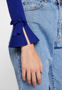 Suéter de punto azul real con mangas acampanadas y detalles de lazo. Combinado con jeans de mezclilla de cintura alta en azul claro. Detalle de textura en la tela visible.