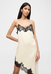 Γυναίκα που φοράει κρεμ σατέν φόρεμα τύπου slip με μαύρη δαντέλα στο ντεκολτέ και ασύμμετρο τελείωμα, στέκεται μπροστά σε απλό φόντο.