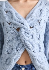 Light blue knitted cardigan featuring a twisted cable design, V-neck, and open front, revealing a midriff. Thick yarn and textured pattern details.