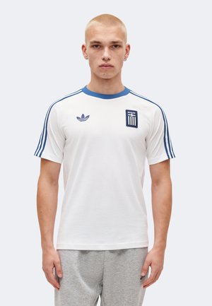 Młody mężczyzna o krótkich blond włosach nosi białą koszulkę Adidas z niebieskimi paskami i greckim emblematem, połączoną z jasnoszarymi spodenkami dresowymi.
