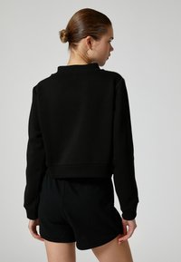 Sort cropped sweatshirt lavet af blødt materiale, med ribbet bund og høj krave, parret med sorte shorts, set fra ryggen.