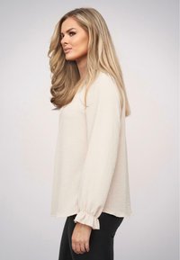 Top a maniche lunghe beige chiaro con una vestibilità rilassata, tessuto testurizzato e polsini con volant. L'orlo è leggermente più lungo sul retro.