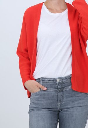 Person trägt eine leuchtend orangefarbene Strickjacke über einem weißen T-Shirt und grauen Jeans, mit einer Hand in der Tasche und der anderen in Kopfnähe erhoben.