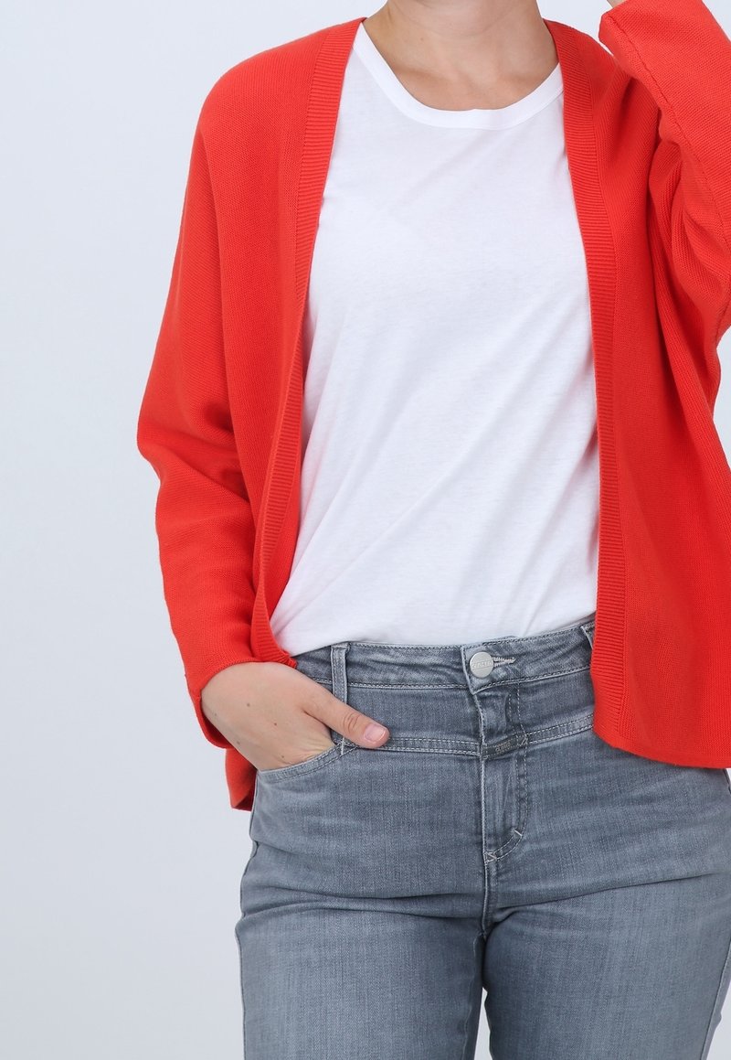 Personne portant un cardigan orange vif par-dessus un t-shirt blanc et un jean gris, une main dans la poche et l'autre levée près de la tête.