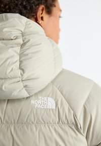 Svijetlosmeđi jakna s punjenjem i velikim kapuljačom, s prišivenim detaljima i bijelim logotipom "The North Face" na ramenu.