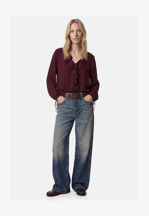 Mujer con una blusa morada oscura con volantes, vaqueros azules descoloridos de pierna ancha, cinturón marrón y zapatos marrones, de pie con las manos en los bolsillos.