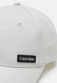 Cappellino da baseball grigio chiaro realizzato in tessuto testurizzato, con un'etichetta nera rettangolare del logo Calvin Klein sulla parte anteriore, design strutturato e corona arrotondata.