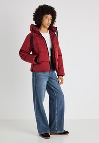 Chaqueta acolchada roja con capucha, detalles en negro y cierre de cremallera, combinada con jeans de pierna ancha azules y zapatos negros sin cordones.