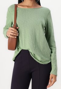 Kvinna iklädd en ljusgrön långärmad topp och svarta figurnära byxor, håller en brun läderaxelbandsväska med sin vänstra hand.