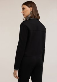 Giacca nera aderente con colletto sartoriale, texture liscia e cuciture sottili sulle spalle, abbinata a un colletto interno grigio.