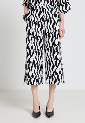 Frau trägt schwarz-weiß gemusterte, weit ausgestellte, kurz geschnittene Hosen mit Wellenmuster und schwarzen spitzen Absätzen, steht mit den Händen an den Seiten.