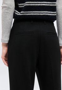 Άτομο που φοράει μαύρο παντελόνι με πίσω τσέπες welt και μαύρο και γκρι ριγέ αμάνικο πουλόβερ πάνω από γκρι μακρυμάνικο πουκάμισο.