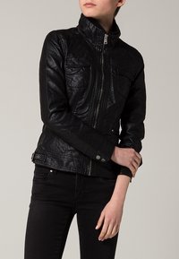 Svart jacka i konstläder med krage, dragkedja framtill och två bröstfickor. Har detaljer med sömmar och knappar med tryckknapp.