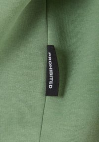 Tissu vert avec une texture lisse. Une étiquette noire indique "PROHIBÉ," cousue sur le côté. Le vêtement est incliné pour montrer à la fois la couleur et l'étiquette.