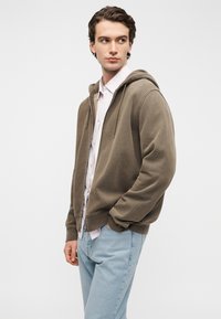 Φούτερ με κουκούλα και φερμουάρ στο μπροστινό μέρος σε ελιά πράσινο, κατασκευασμένο από απαλό ύφασμα. Διαθέτει άνετη εφαρμογή, ριμπ μανσέτες και τσέπες καγκουρό. Συνδυάζεται με ανοιχτό ροζ πουκάμισο.