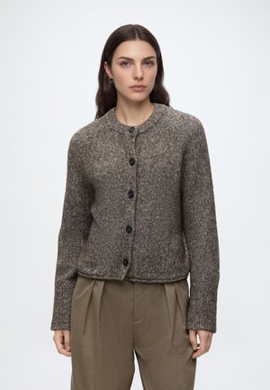 Donna con lunghi capelli scuri che indossa un cardigan marrone di maglia abbottonato e pantaloni marroni larghi, vista frontale su sfondo bianco.