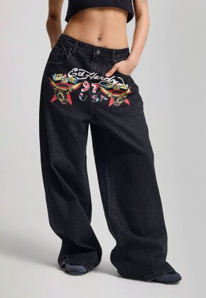 Sorte vidde jeans med farverig tyr og stjerne grafik, 'Ed Hardy 97 USA' tekst, båret med cropped sort top og synlig navlepiercing.
