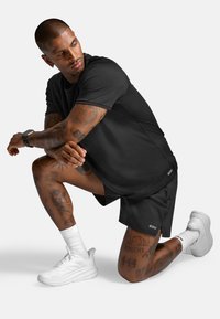 Czarna sportowa koszulka i spodenki noszone przez osobę klęczącą. Na ramionach i nogach widoczne tatuaże, do tego białe trampki i skarpetki.