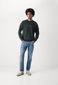 Donkergroene sweatshirt met ronde hals en wit logo, gecombineerd met lichtblauwe jeans en sneakers met groene accenten. Glad textuur van de stof.