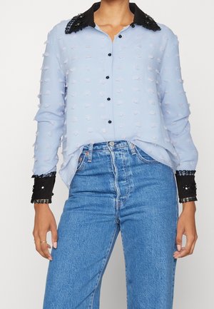 Hellblaue Bluse mit strukturierten Punkten, schwarzem Spitzenkragen und Manschetten, in hoch taillierte blaue Jeans gesteckt getragen.