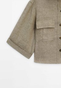 Chemise en lin beige à manches courtes avec fermeture par boutons et grande poche à rabat sur la poitrine, présentée sur fond blanc.
