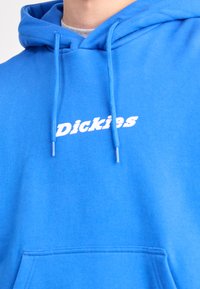 Sudadera azul hecha de una tela suave, con un bolsillo frontal, cordones y el logo "Dickies" en blanco en el pecho.