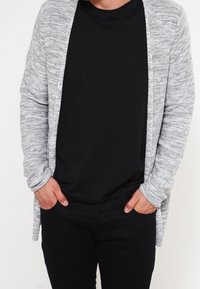 Grå, texturerad cardigan med öppen front, som bärs över en svart t-shirt och svarta byxor. Cardiganen har långa ärmar och en avslappnad passform.