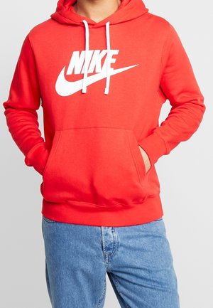 Person trägt einen leuchtend roten Nike-Hoodie mit weißen Kordeln und Logo, Hände in der Vorderseite-Tasche, kombiniert mit hellblauen Jeans.