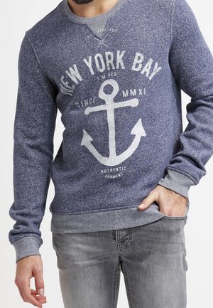 Pull en coton bleu marine avec une texture mouchetée, présentant un graphique d'ancre blanche et un texte imprimé à l'avant. Poignets et ourlet côtelés.