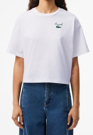 T-shirt blanc court en coton, avec des manches courtes et un logo brodé vert sur la poitrine. Assorti à un jean taille haute.