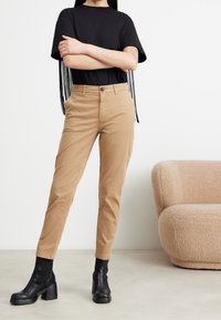 Svart kortärmad t-shirt med fransdetaljer, i kombination med beiga tajta byxor. Modellen bär svarta klackboots. Neutral bakgrund.