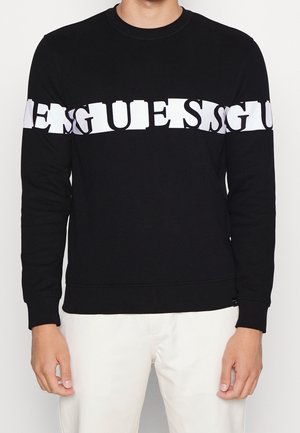 Mann trägt ein schwarzes Langarmshirt mit Rundhalsausschnitt und einem weißen, verzerrten horizontalen Textband über der Brust und den Oberarmen.