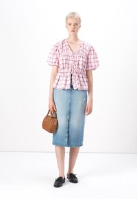 Blusa a quadri rosa e bianchi con maniche a sbuffo e nodo frontale, abbinata a una gonna in denim blu di lunghezza media. Tiene una piccola borsa marrone.