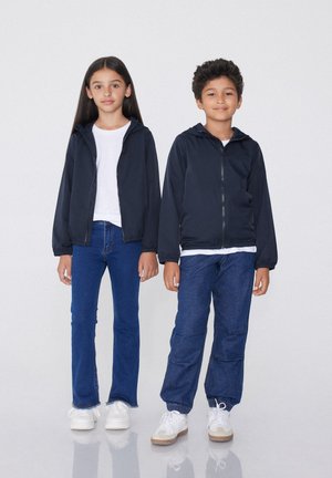 Deux enfants se tiennent côte à côte, portant des coupe-vent navy avec capuches, des chemises blanches et des jeans bleus. Le pantalon du garçon a des poignets élastiques et des poches.