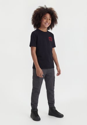 Enfant souriant aux cheveux bouclés, portant un t-shirt graphique noir, un pantalon cargo gris foncé et des baskets montantes noires, debout sur un fond blanc.