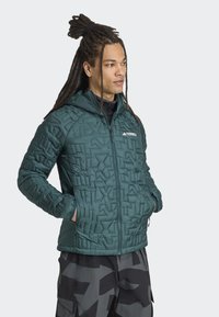 Teal gewatteerde jas met capuchon, voorzien van een geometrisch patroon en ritsluiting. Logo op de borst, zijzakken en een relaxte pasvorm.