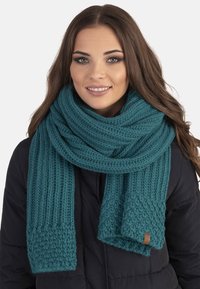 Écharpe en tricot turquoise avec un motif texturé et des extrémités côtelées. Fabriquée en matériau doux avec une petite étiquette en cuir.